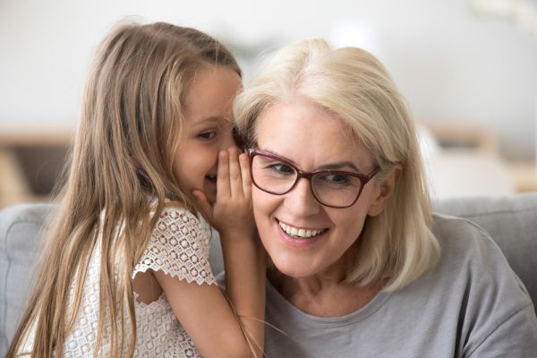 A heartwarming moment between a young granddaughter whispering into her grandmother's ear, both sharing a joyful, loving smile.