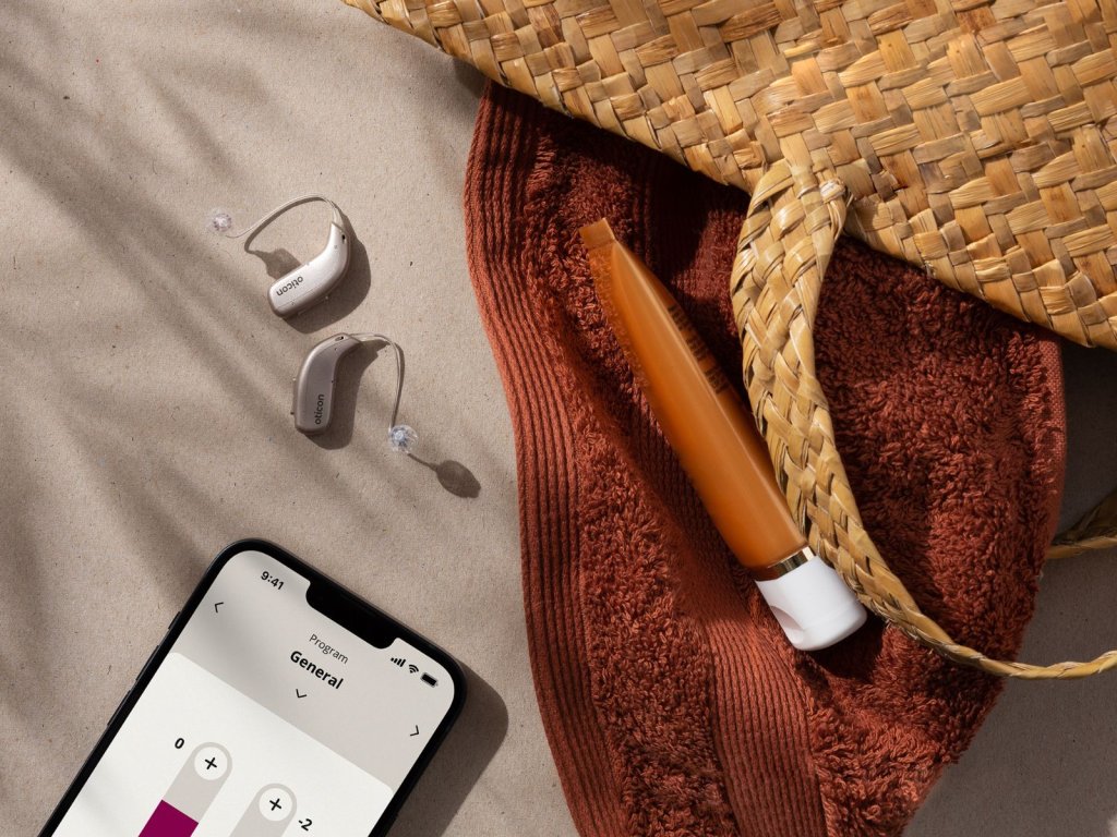 Oticon Real hearing aids and a phone with the Oticon app on beach sands.