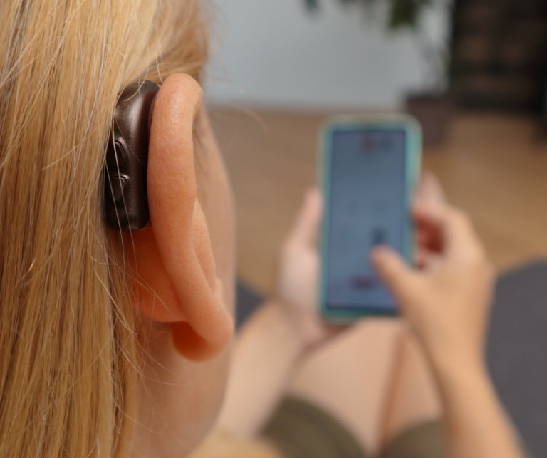 Close up of blonde woman wearing hearing aid on ear and pressing button while using smartphone at home.