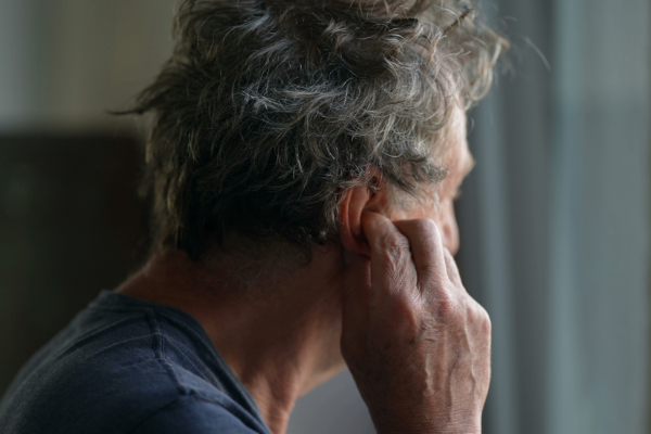 Older man touching and scratching ear standing by window.