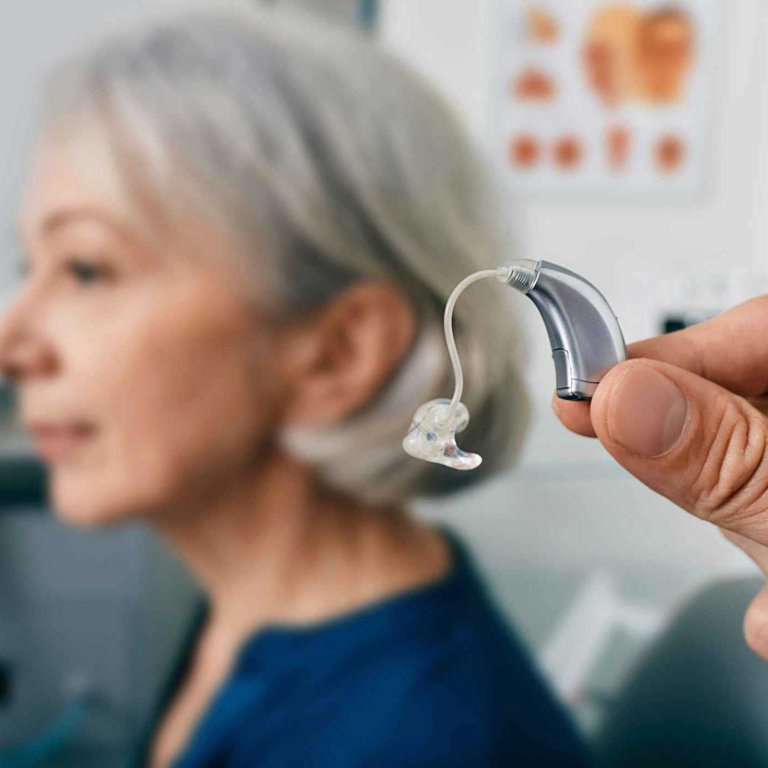 Close-up of a hand holding a hearing aid in the foreground, with a woman in the background.