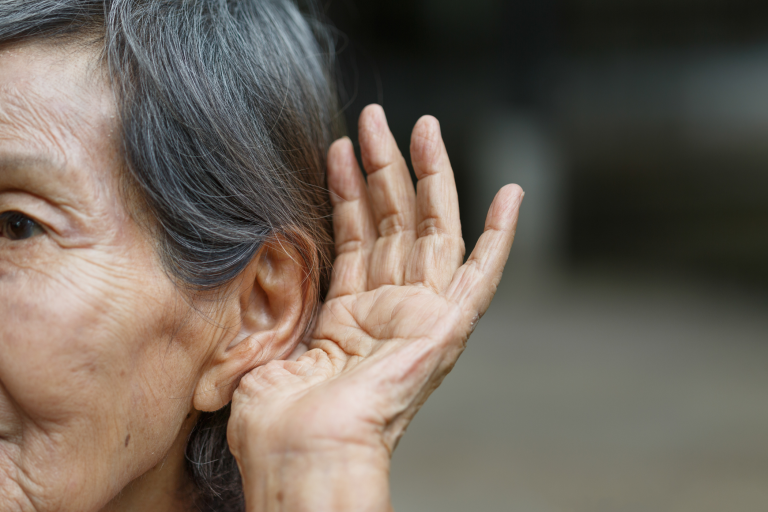 Older woman with wrinkles and gray hair, cupping her hands behind her ears as if straining to hear.